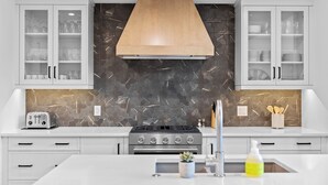 A modern kitchen with white cabinetry, a central island, a built-in stove, and a backsplash with a geometric pattern. - Spring Creek Vacations 4 BR Villa TMV18 (Canmore)