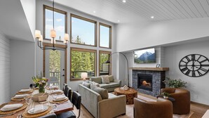 A modern living room with a fireplace, large windows, and a dining area set up for a meal. - Spring Creek Vacations 4 BR Villa TMV18 (Canmore)