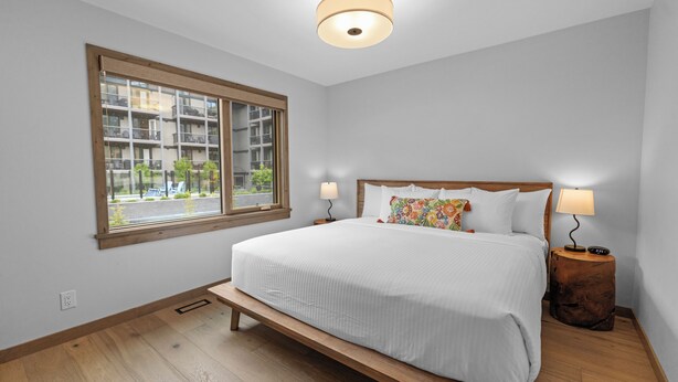 A bedroom with a large bed, two bedside lamps, a wooden bench, a window with a view of buildings, and a ceiling light fixture. - Spring Creek Vacations 4 BR Villa TMV18 (Canmore)