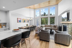A modern kitchen with a marble island, dining area with a wooden table, and a living room with a fireplace and large windows. - Spring Creek Vacations 4 BR Villa TMV24 (Canmore)