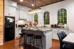 A modern kitchen with white cabinetry, a black refrigerator, a central island with stools, and large windows offering views of the outdoors. - Romantic Cabin Getaway Sauna, Hot tub & Pizza Oven (Broken Bow)
