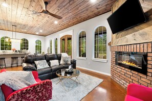 A modern living room with a fireplace, leather sofas, and a kitchen in the background. - Romantic Cabin Getaway Sauna, Hot tub & Pizza Oven (Broken Bow)