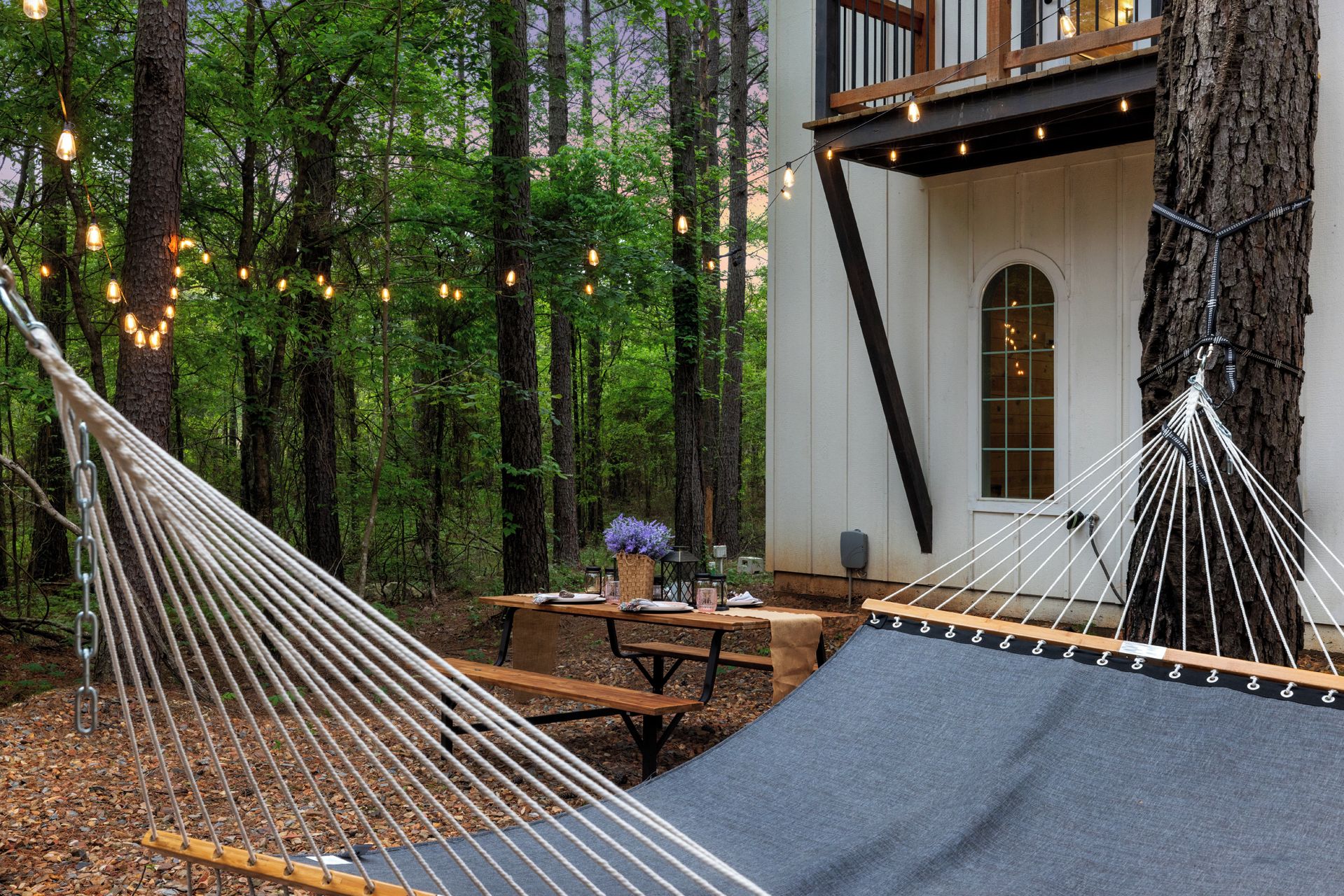 A hammock set up in a forested area with a wooden table and benches nearby, suggesting a relaxing outdoor space.