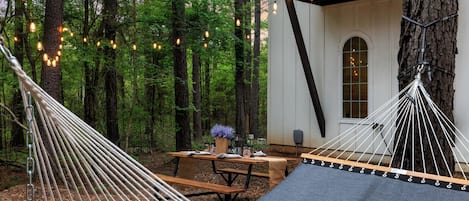 A hammock set up in a forested area with a wooden table and benches nearby, suggesting a relaxing outdoor space.