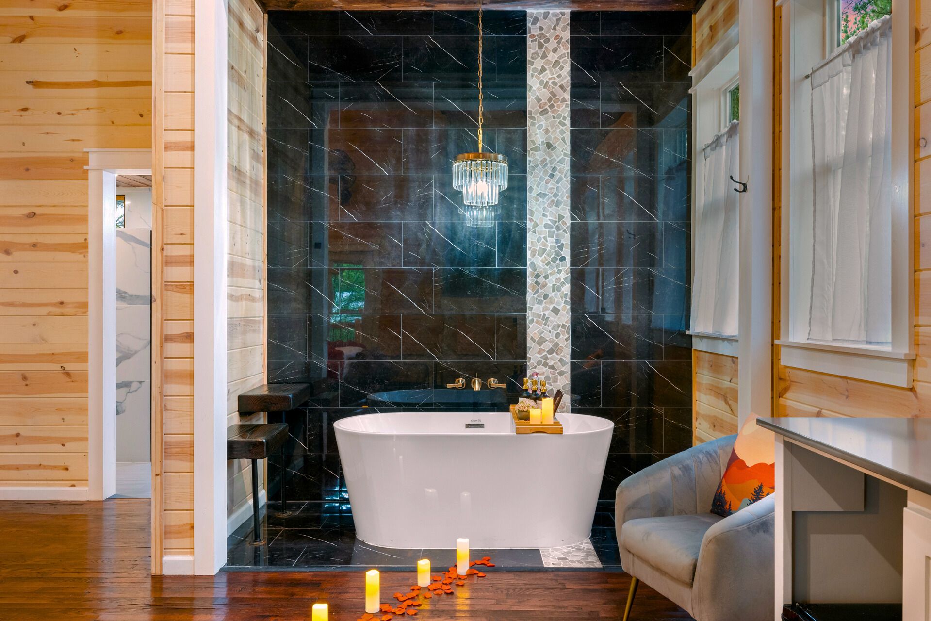 A modern bathroom with a freestanding bathtub, a chandelier, and a seating area with a cushion.