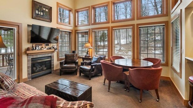 A spacious living room with a fireplace, a flat-screen TV, a dining area with a round table and chairs, and large windows with blinds. - Slopeside Boyne Mountain Retreat (Boyne Falls)