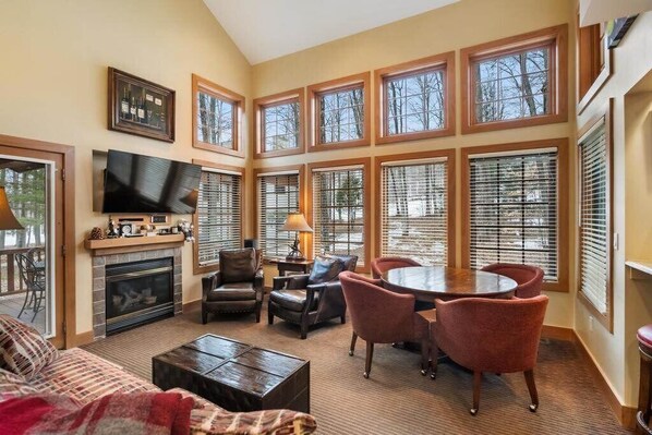 A spacious living room with a fireplace, a flat-screen TV, a dining area with a round table and chairs, and large windows with blinds.