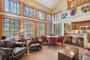A spacious living area with large windows, a dining table with chairs, a fireplace, and a kitchen area.