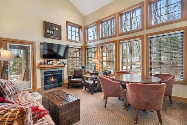 A spacious living room with a fireplace, comfortable seating, a television, and large windows offering a view of the outdoors.