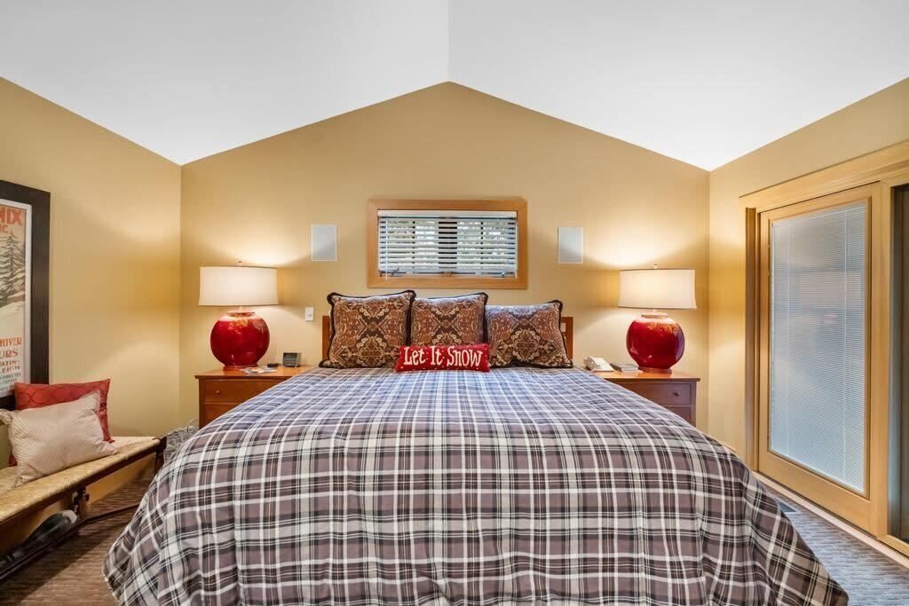 A bedroom with a large bed, two bedside lamps, a window with blinds, and a decorative pillow with the text 'Let It Snow'.