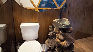 Un baño con pared de madera, inodoro blanco, un cesto de basura azul y una decoración de rocas.