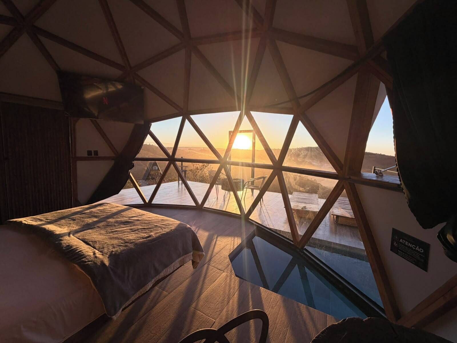 A room with a bed, a glass table, and a view of the sunset through a geometric window.