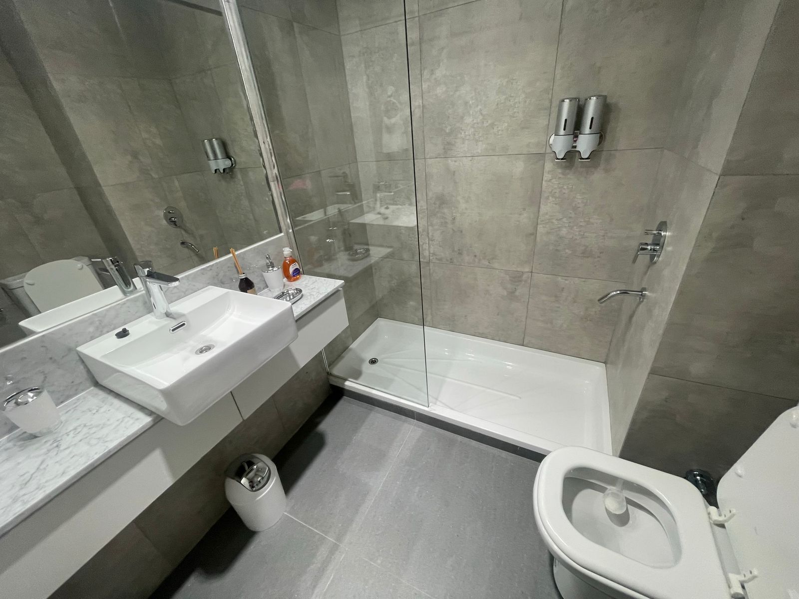 A modern bathroom with a large mirror, two sinks, a toilet, and a shower area.