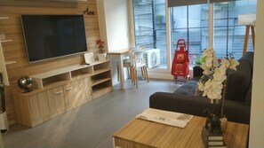 Una sala de estar moderna con un televisor de pantalla plana, un mueble de madera para entretenimiento, un sofá negro, una mesa de centro con un libro y un jarrón de flores, y un bolso rojo en el suelo.