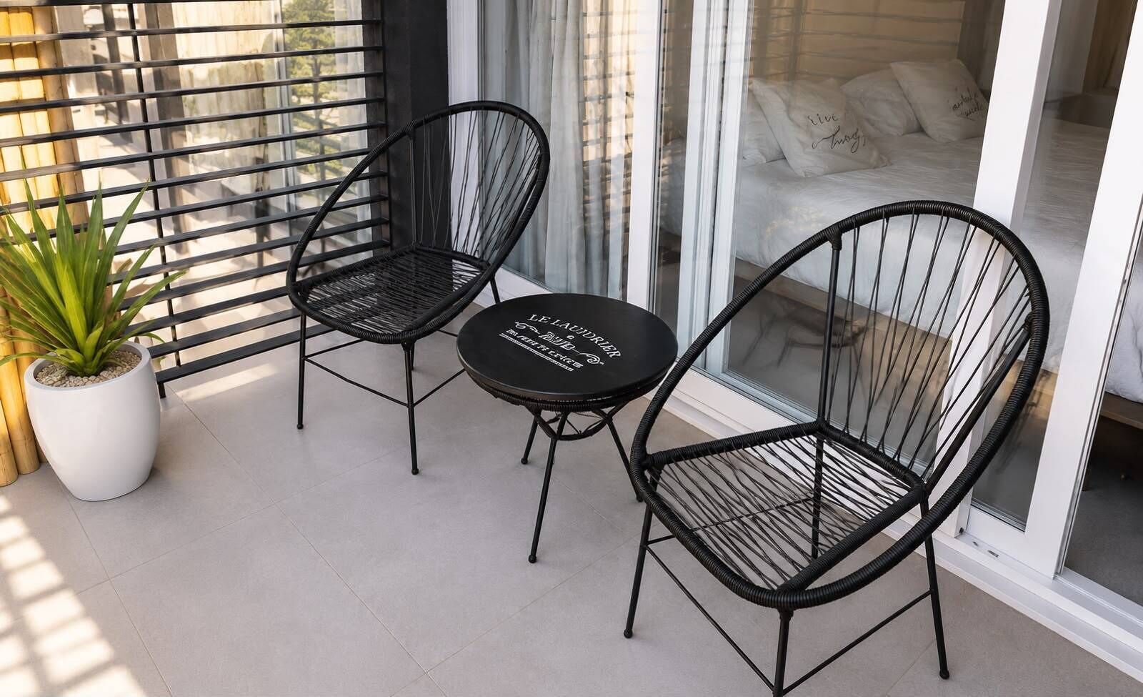 A balcony with two black wicker chairs and a small round table, a potted plant, and a bed with white bedding and pillows.