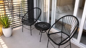 En balkon med to sorte fletstole, et rundt bord, en potteplante samt en seng med hvide sengetæppe og puder.