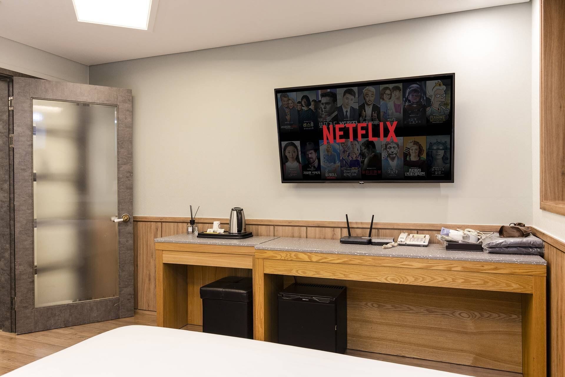 Quarto de hotel com uma secretária de madeira, uma televisão fixada na parede exibindo a interface da Netflix e uma porta com painéis de vidro jateado.