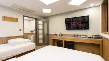 Quarto de hotel com uma cama, uma escrivaninha com um computador, uma televisão na parede e uma porta que dá para outro quarto.