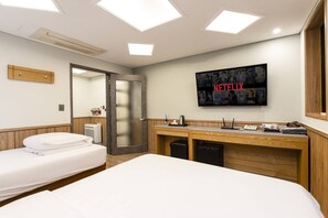 Hotellihuone, jossa on sänky, työpöytä tietokoneella, seinälle asennettu televisio ja ovi, joka johtaa toiseen huoneeseen.
