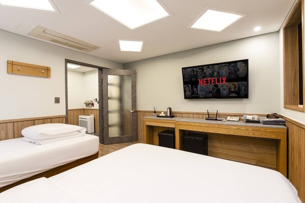 Quarto de hotel com uma cama, uma escrivaninha com um computador, uma televisão na parede e uma porta que dá para outro quarto.
