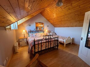 A bedroom with a sloped ceiling, two beds, a bedside table with lamps, a small bedside table, and a wooden chair.