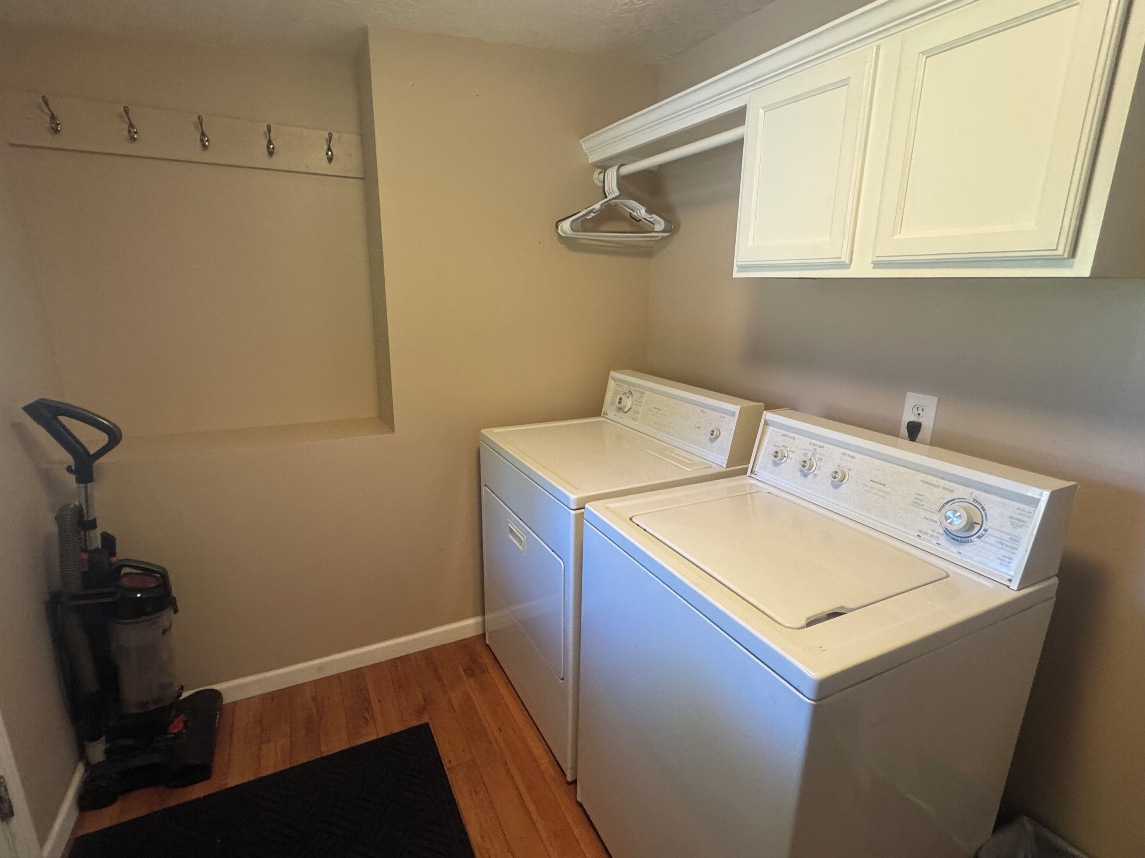 A laundry room with two white front-loading washers and dryers, a vacuum cleaner, and a clothes drying rack.