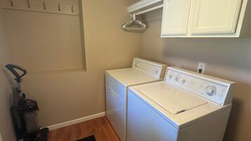 A laundry room with two white front-loading washers and dryers, a vacuum cleaner, and a clothes drying rack.
