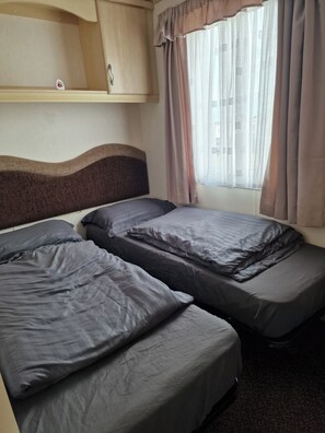 Une chambre avec deux lits, une tête de lit, une fenêtre avec des rideaux et une étagère ornée d’un objet décoratif.