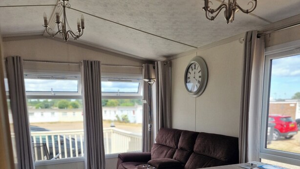 A compact living space with a sofa, a small table, a clock, and a chandelier. - Caravan Sleeps 6 with Parking (Great Yarmouth)