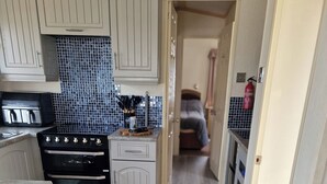 A compact kitchen with white cabinetry, a black stove, and a blue-tiled backsplash. - Caravan Sleeps 6 with Parking (Great Yarmouth)