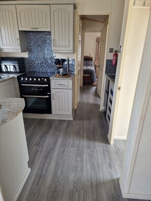 A compact kitchen with white cabinetry, a black stove, and a blue-tiled backsplash. - Caravan Sleeps 6 with Parking (Great Yarmouth)