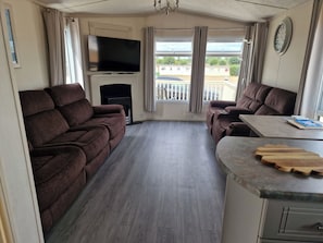 A living room with a flat-screen TV, two sofas, a small table, and a kitchen area with a wooden countertop. - Caravan Sleeps 6 with Parking (Great Yarmouth)
