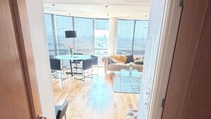Een ruime woonkamer met een hoge plafond, houten vloer en grote ramen met uitzicht op de stad.