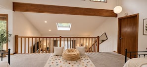 Un dormitorio con cama, alfombra de lunares, sillón de mimbre, techo de madera y tragaluz.