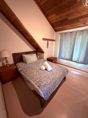 Ein Schlafzimmer mit schrägen Wänden, einem Bett mit gemusterten Betttextilien, einem Nachttisch mit Lampe und einem Fenster mit Vorhängen.
