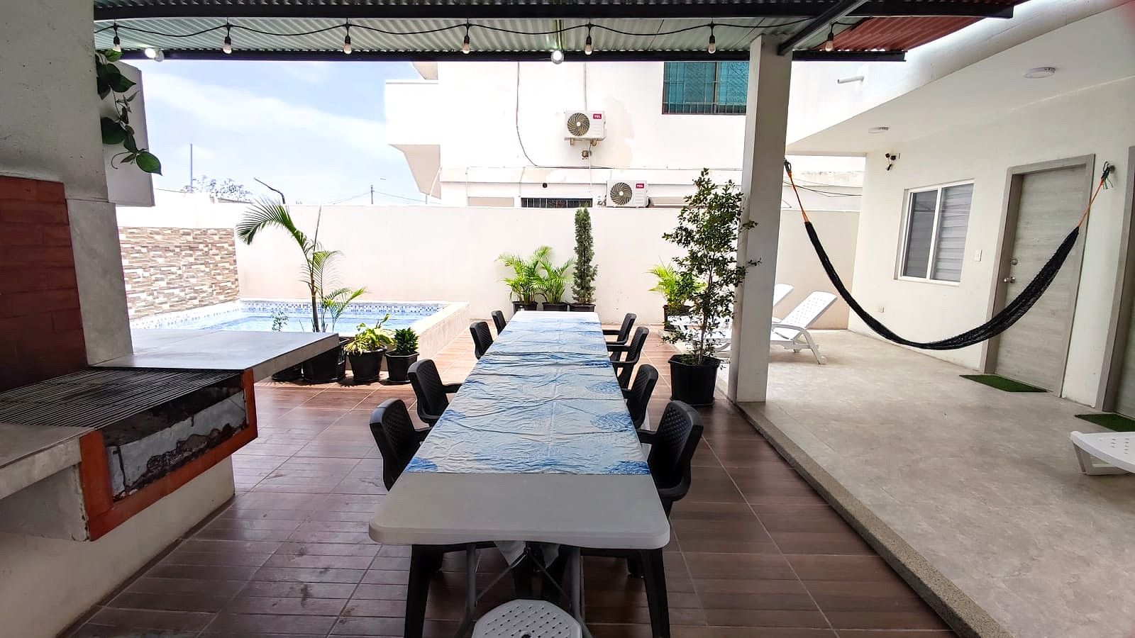 Um pátio externo com uma mesa comprida e cadeiras, rodeado por vasos de plantas e uma piscina.
