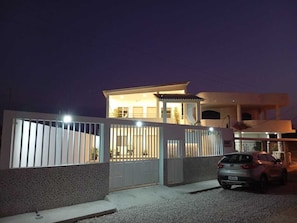 A modern two-story house with a gated entrance, a car parked in the driveway, and a well-lit exterior at night.