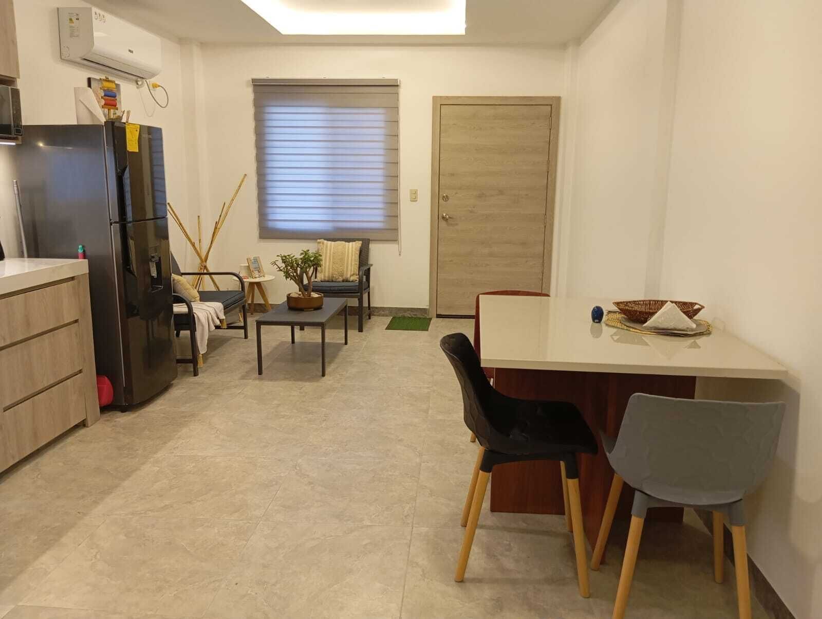 Eine kleine Küche mit einem Kühlschrank, einem Esstisch mit Stühlen und einem Fenster mit Jalousien.