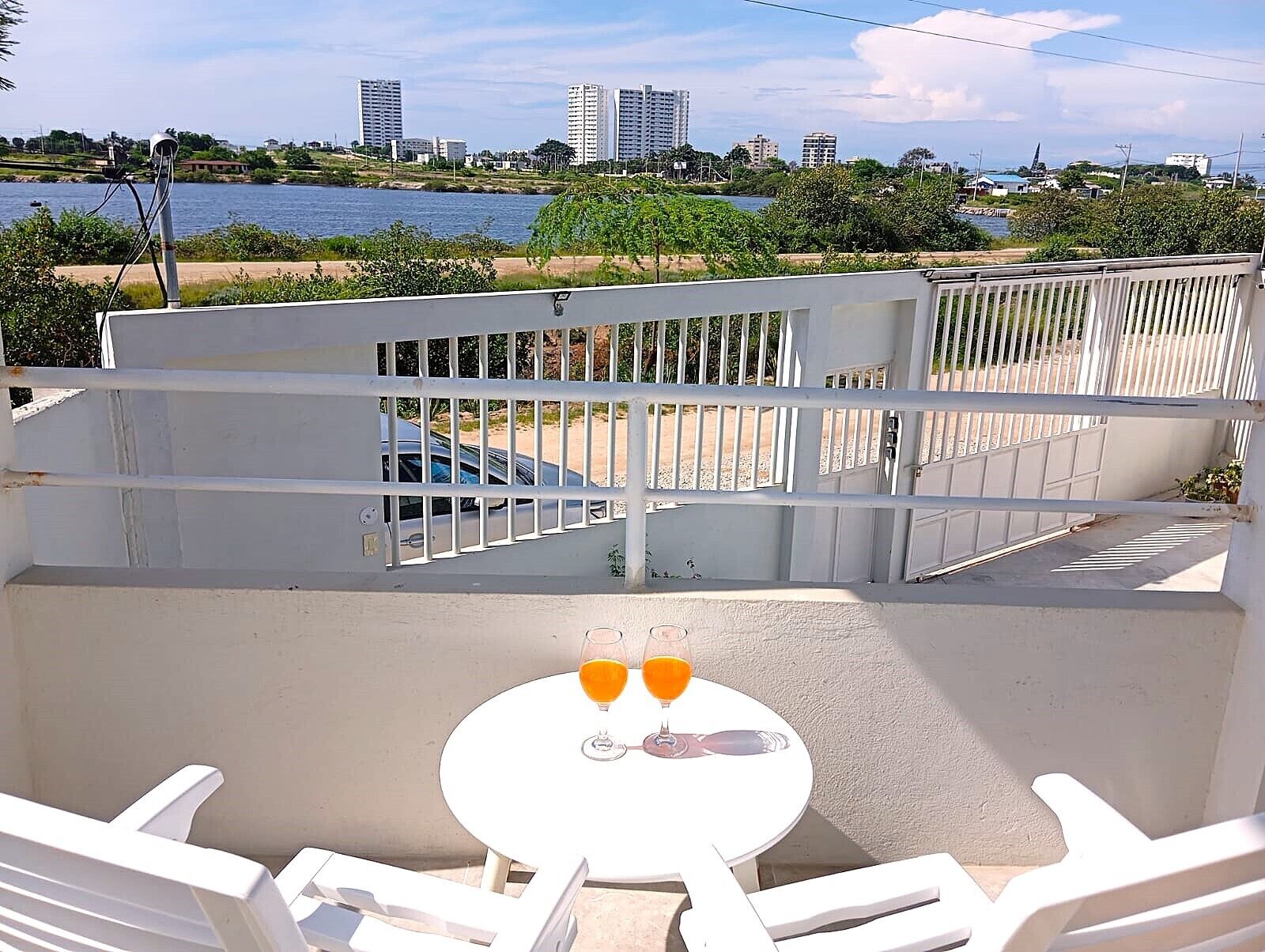 A balcony with a white railing, a table with two glasses of orange juice, and a view of a cityscape and water.