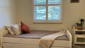 Un lit simple avec une tête de lit, une fenêtre au-dessus, une table de chevet avec une lampe et une petite étagère avec une plante.