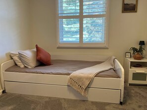A single bed with a headboard, a window above it, a bedside table with a lamp, and a small shelf with a plant.
