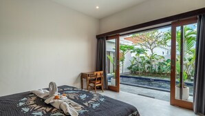 A hotel room with a bed, a small table, and a view of a garden through a sliding glass door. - Sona Villa Bali (Badung)