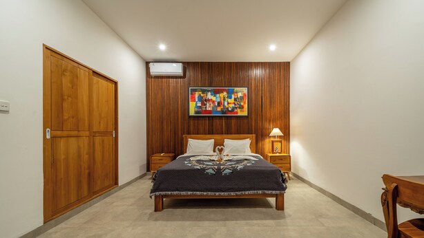 A bedroom with a wooden bed, a nightstand, a bedside table, a wall-mounted air conditioner, and a colorful abstract painting. - Sona Villa Bali (Badung)