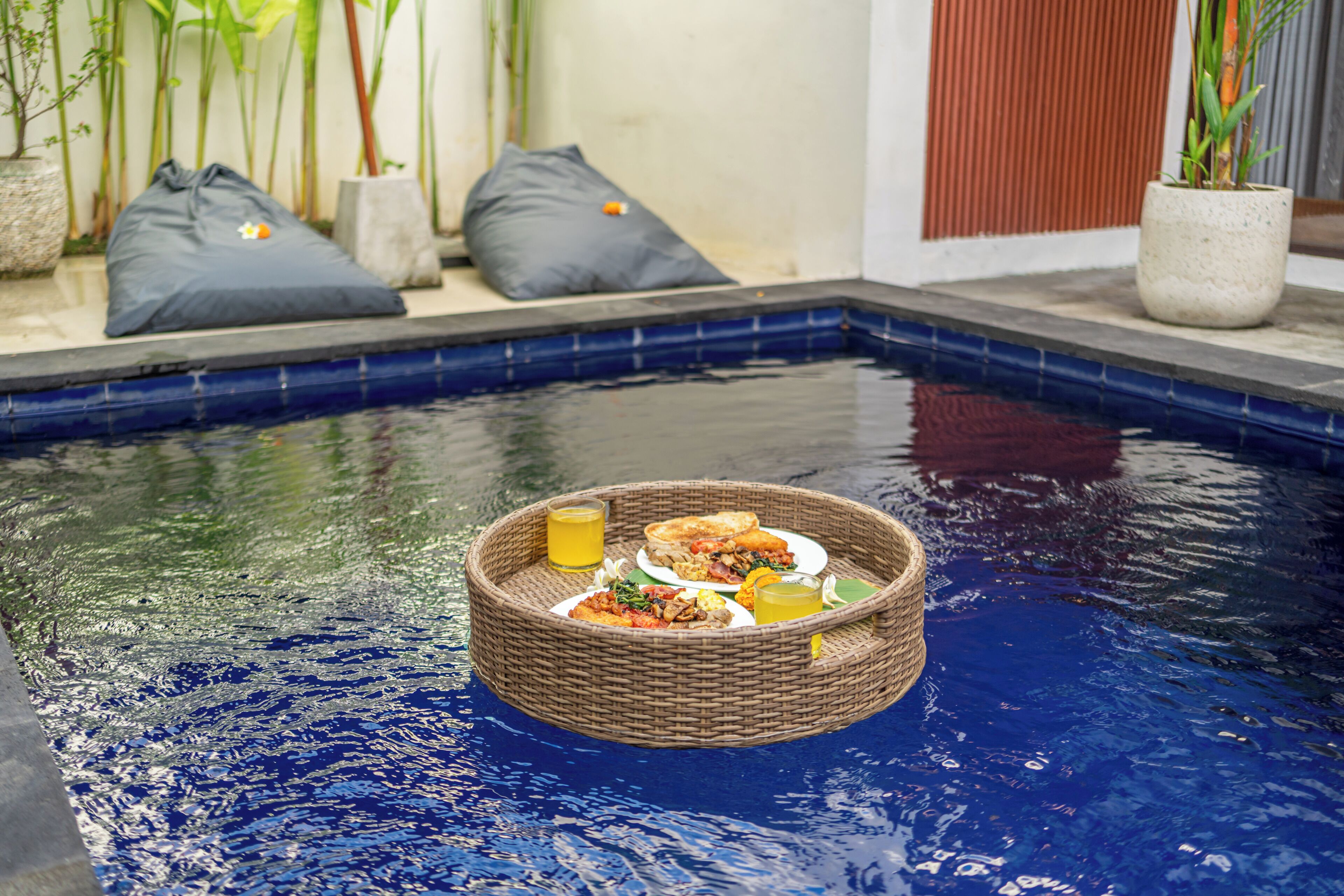 Une table dressée avec un panier placé au bord d’une piscine, entourée de coussins et de plantes.