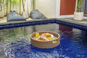Una zona de comedor junto a una piscina, con una cesta en el borde, cojines y plantas alrededor.