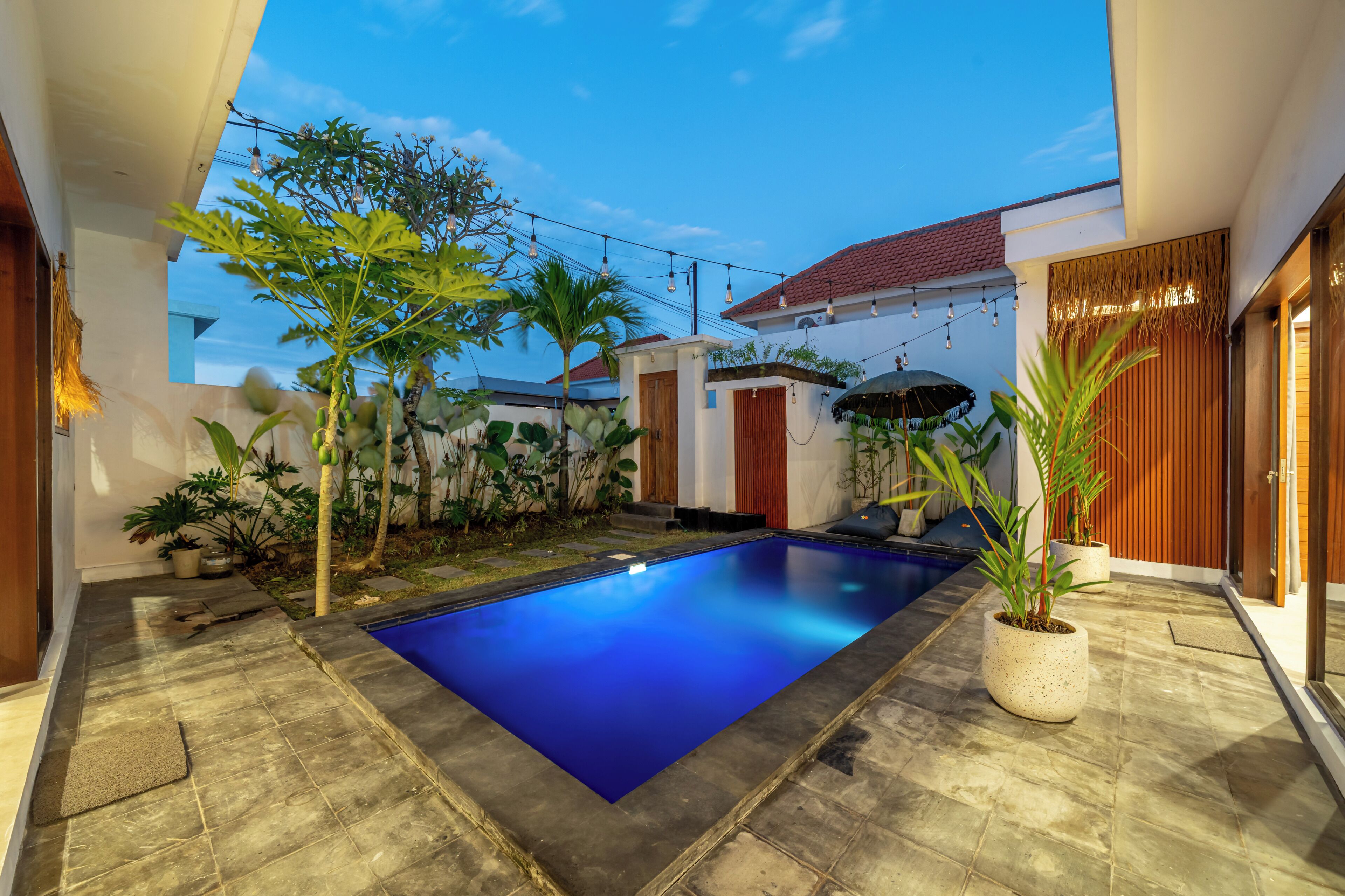Una piscina con plataforma de azulejos, rodeada de plantas en macetas y un pequeño jardín.