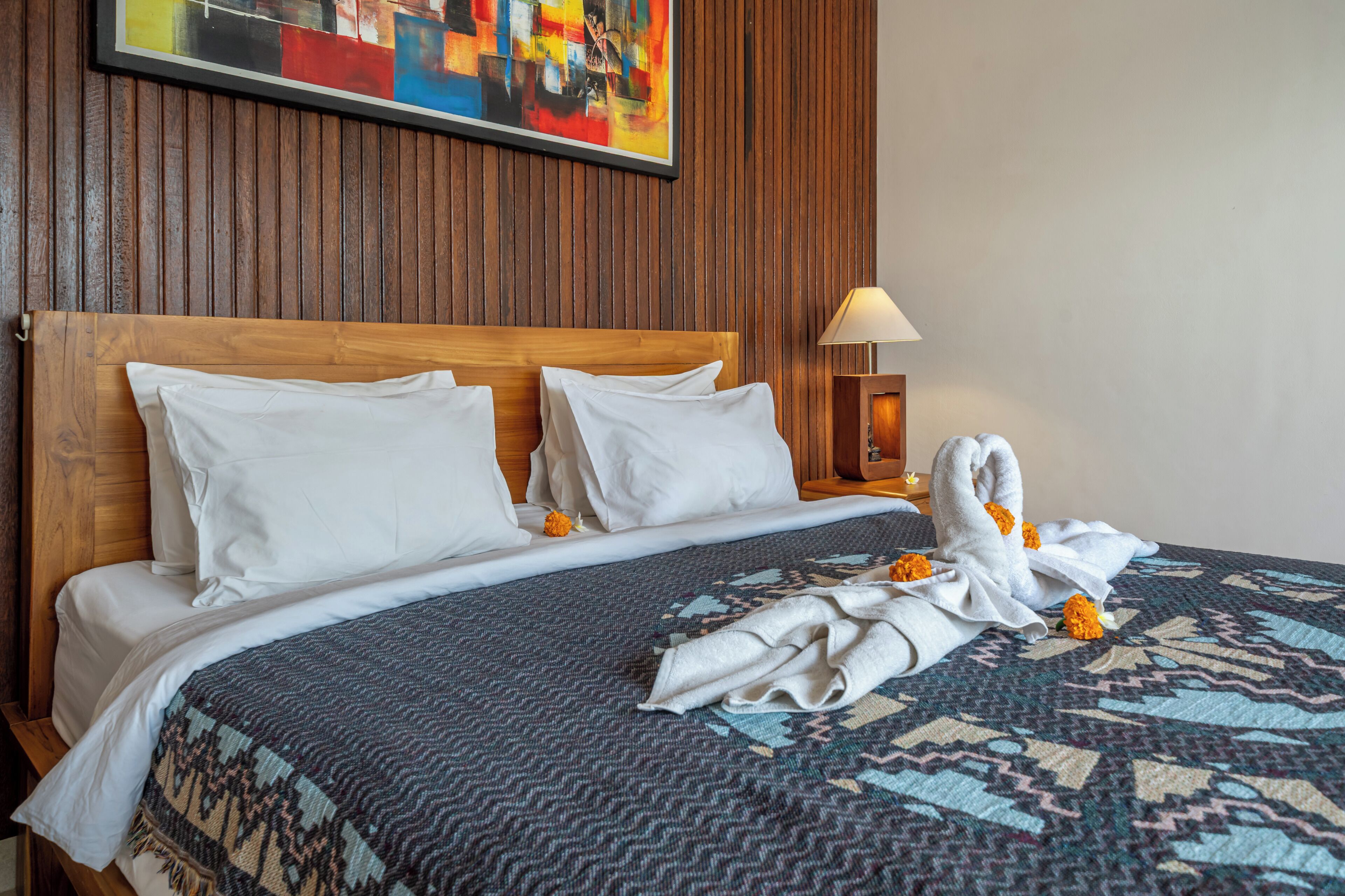 Habitación de hotel con cabecera de madera, edredón estampado, sábanas blancas, un cuadro en la pared y una lámpara de noche.