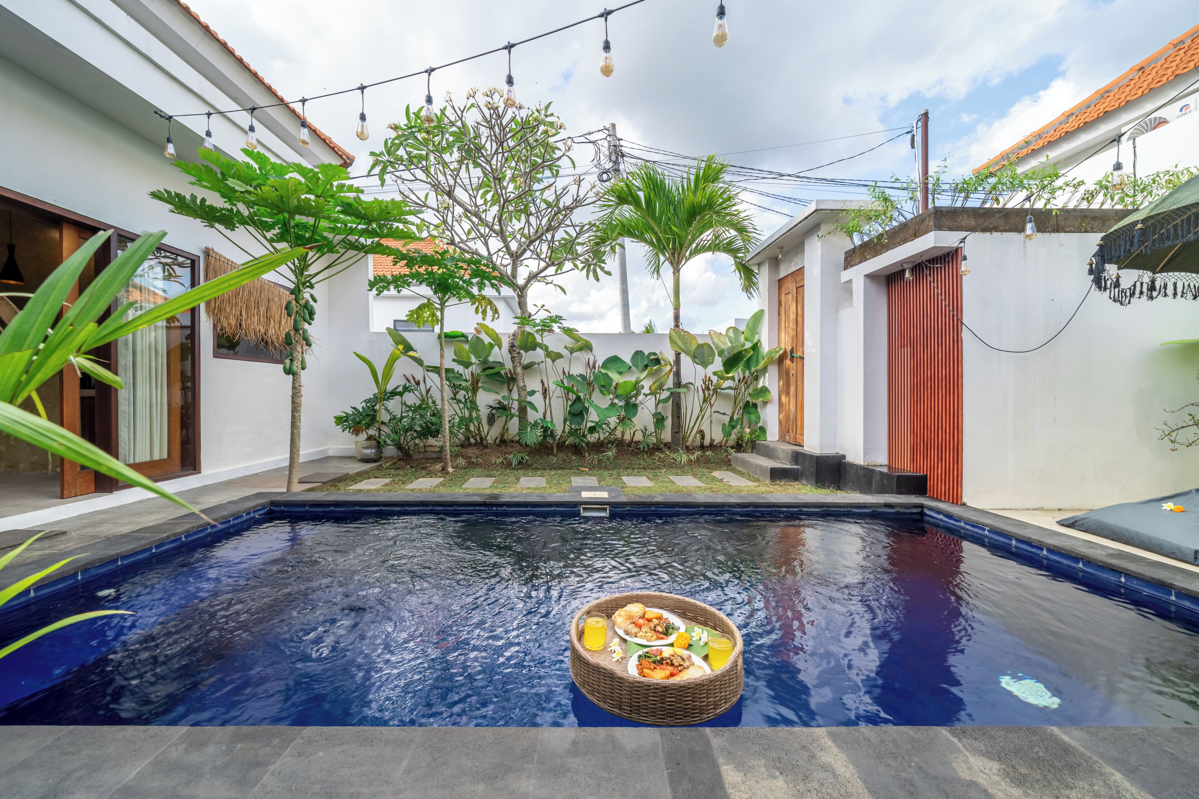 A swimming pool with a floating table of food and drinks, surrounded by lush greenery and a white building with a red door.