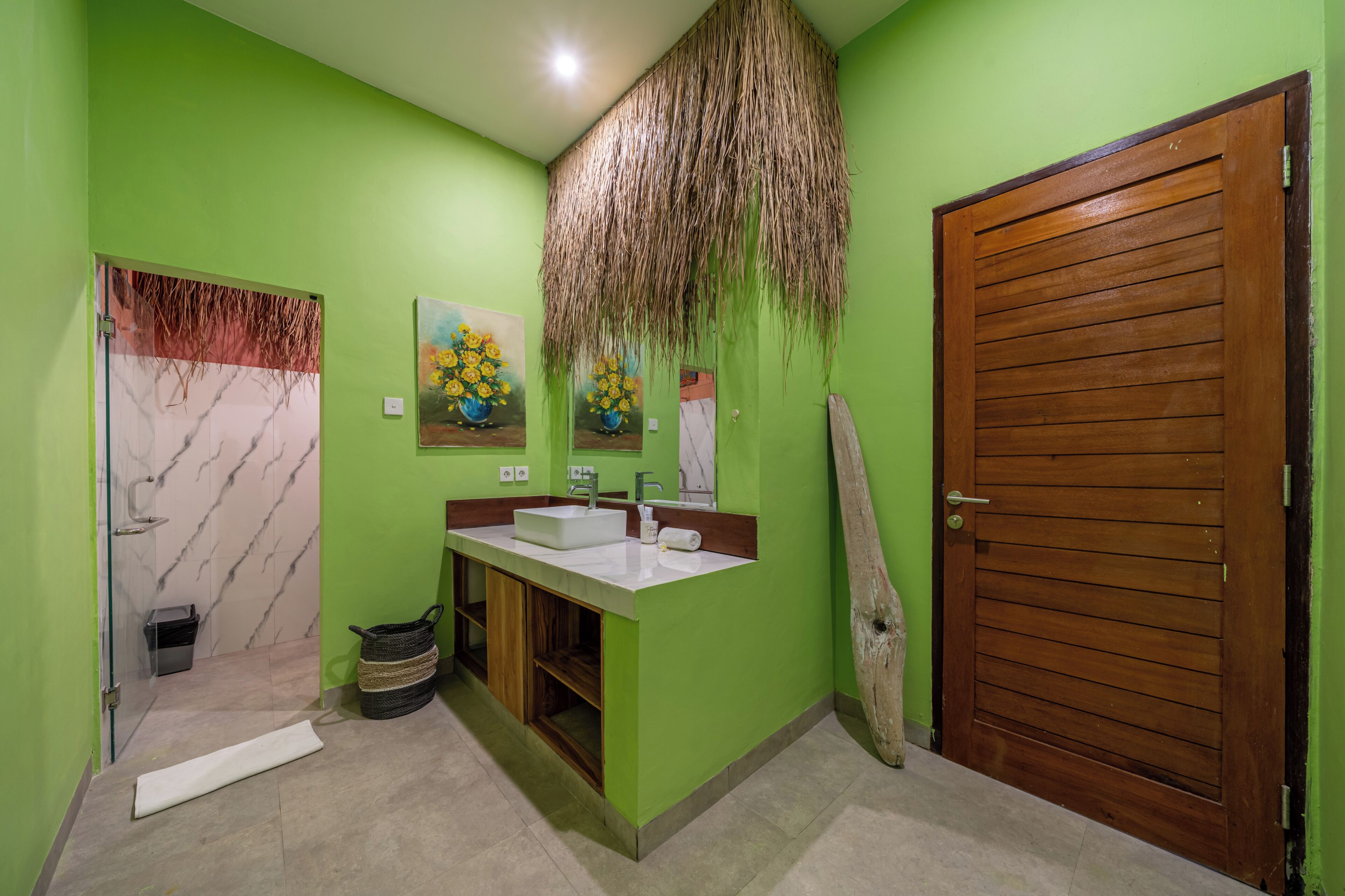 Un baño con pared verde, puerta de madera, lavabo de mármol y un cuadro de flores amarillas.