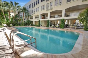 Una piscina con barandilla metálica de seguridad, rodeada de palmeras y un edificio de varios pisos con balcones.